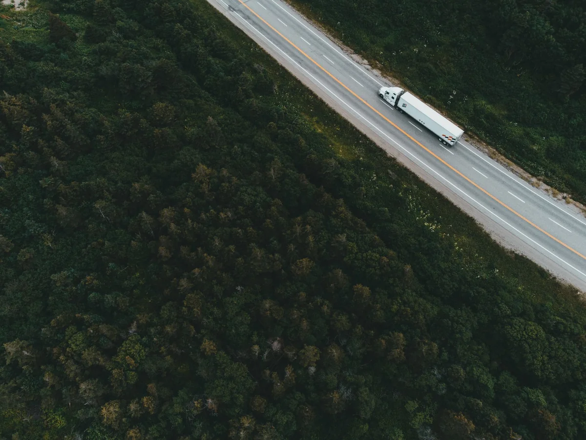 Real aerial reference photograph of a white container truck on a highway bordered by forest.